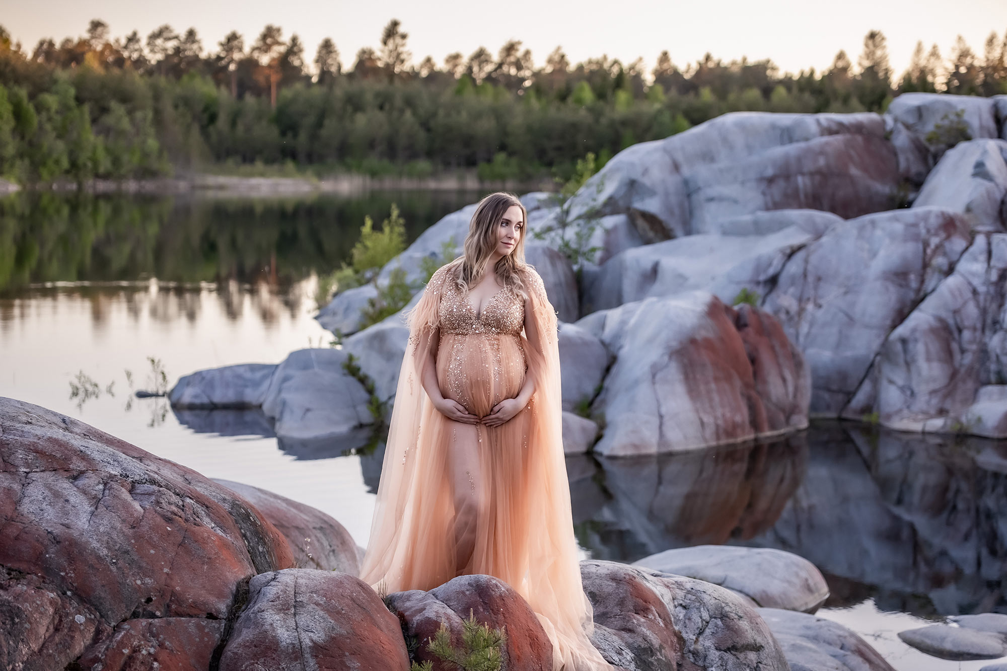 Gravidfotografering vid havet på en strand om våren. I bakgrunden syns klippor och stilla ljus över skärgården.