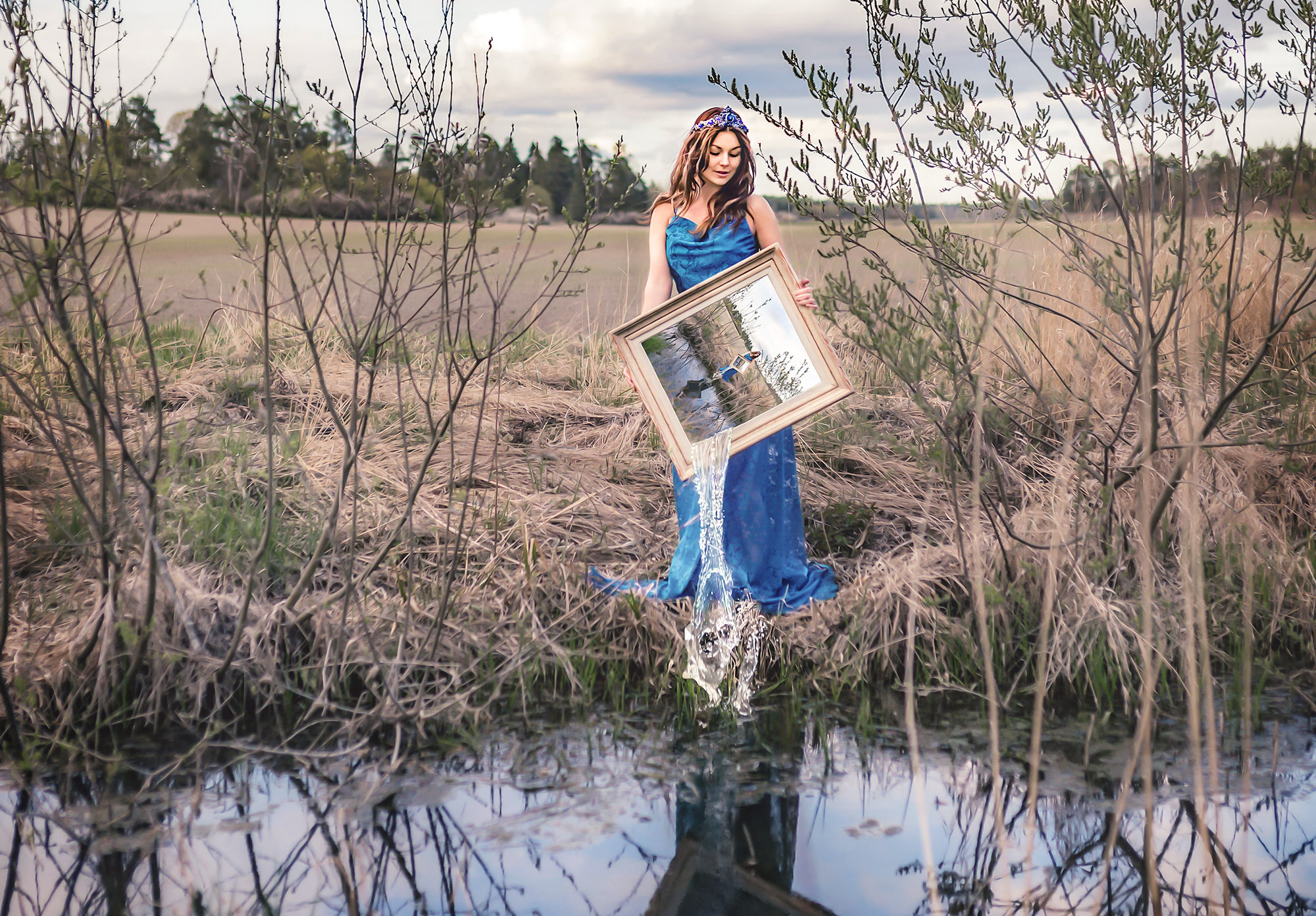 En bild med en kvinna som står vid en sjö och håller i en tavla med bilden av kvinnan. Hon häller ut vatten från tavlan ner i sjön.
