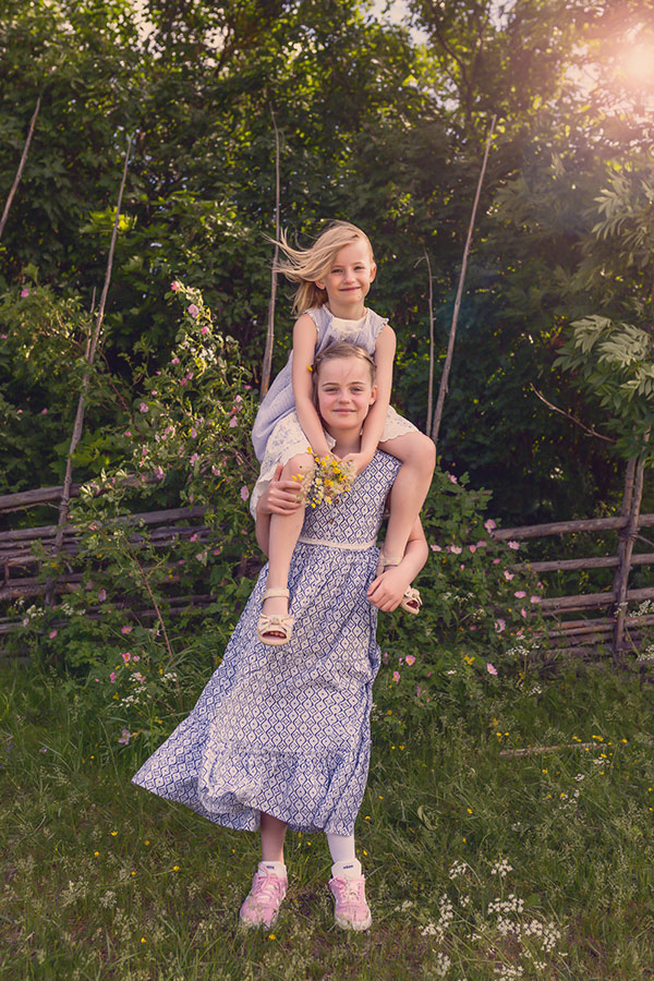 Två systrar i grönskande sommar. Storasyster bär lillasyster på axlarna.