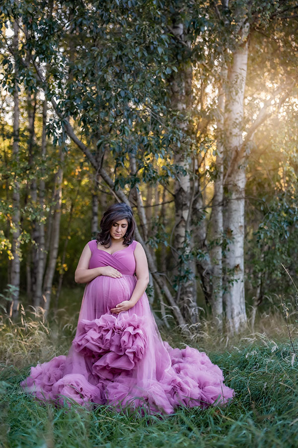 En gravid kvinna i rosa tyllklänning står i högt gräs vid grönskande björkar. I bakgrunden letar sig kvällssolen in.
