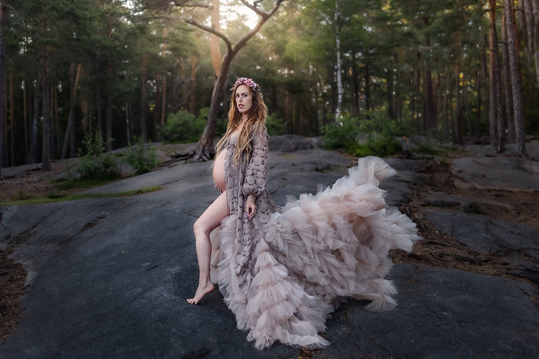 Gravidfotografering i en skog med klippor. Ljuset strilar in genom grenarna och lyser på den gravida kvinnan som har på sig en flygande beige tyllklänning som är öppen för magen.