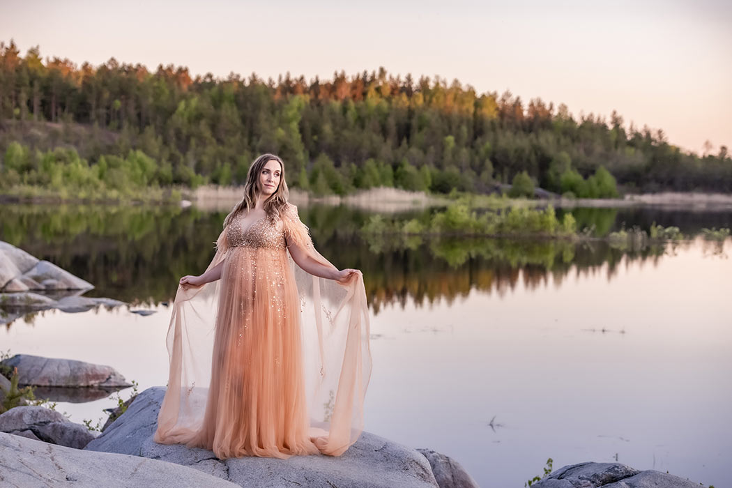 Gravidfotografering i fantastiskt kvällsljus. Det spegelblanka vattnet speglar skogen i bakgrunden och den gravida kvinnan har en vacker gravidklänning.