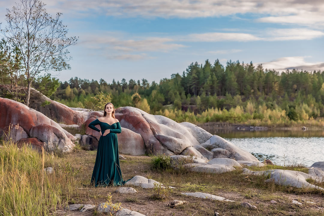 En gravid kvinna i grön gravidklänning står på en stenig strand bland klippor och höga gräs. Kvinnan håller om sin mage och tittar in i kameran. I bakgrunden syns en grönskande skog som börjat gulna i hösten.