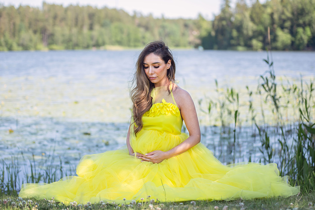 Gravidfotografering vid en sjö. Den gravida kvinnan har på sig en gul tyllklänning och sitter på en gräsmatta framför en sjö med näckrosor.