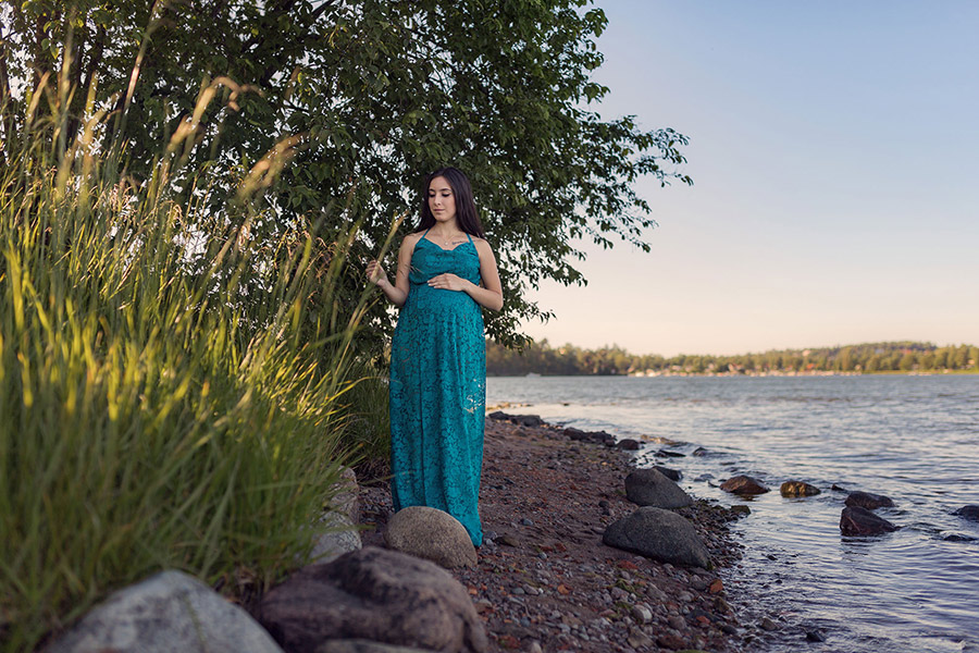 En gravid kvinna i blå spetsklänning står på en smal stenstrand bredvid vid höga grässtrån och grönskande träd. I bakgrunden syns vattnet och andra sidan av sjön.