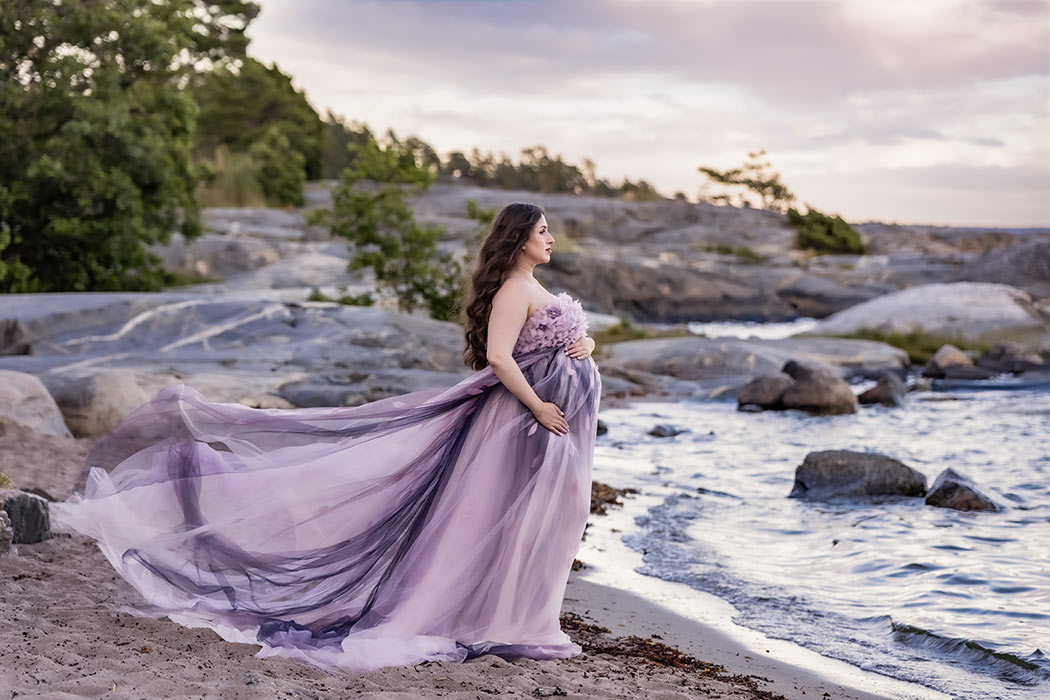 En vacker kvinna står på en stormande strand med klippor i bakgrunden. Kvinnan är gravid och hennes extravaganta lila klänningar blåser i vinden.