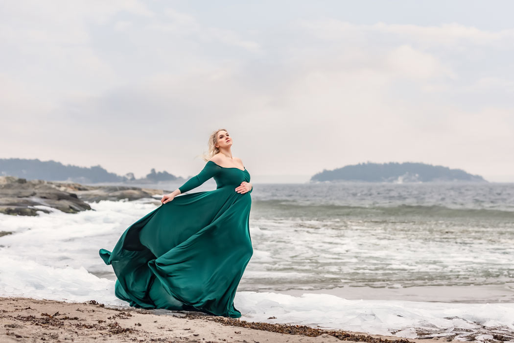 En dramatiskt vacker bild från en gravidfotografering vid havet om vintern. Den gravida kvinnan har på sig grön klänning som flyger i vinden.