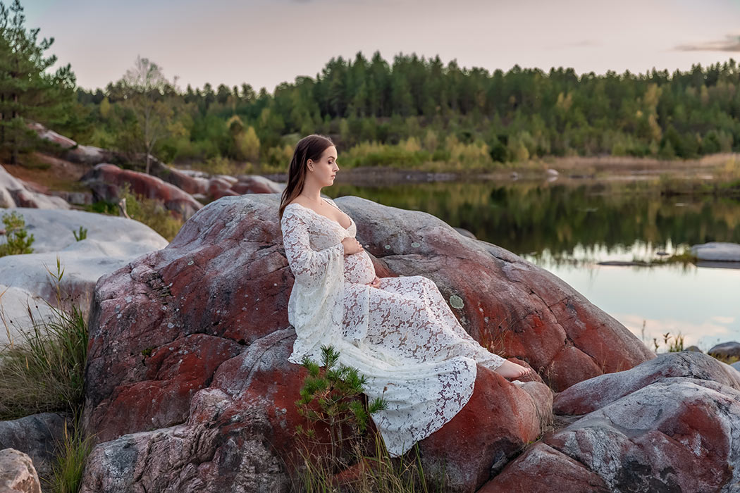 Gravidfotografering bland klippor i ett stenbrott. Det gravida kvinnan sitter på en stor sten och har på sig en vit spetsklänning. I bakgrunden syns en spegelblank sjö och grönskande skog.