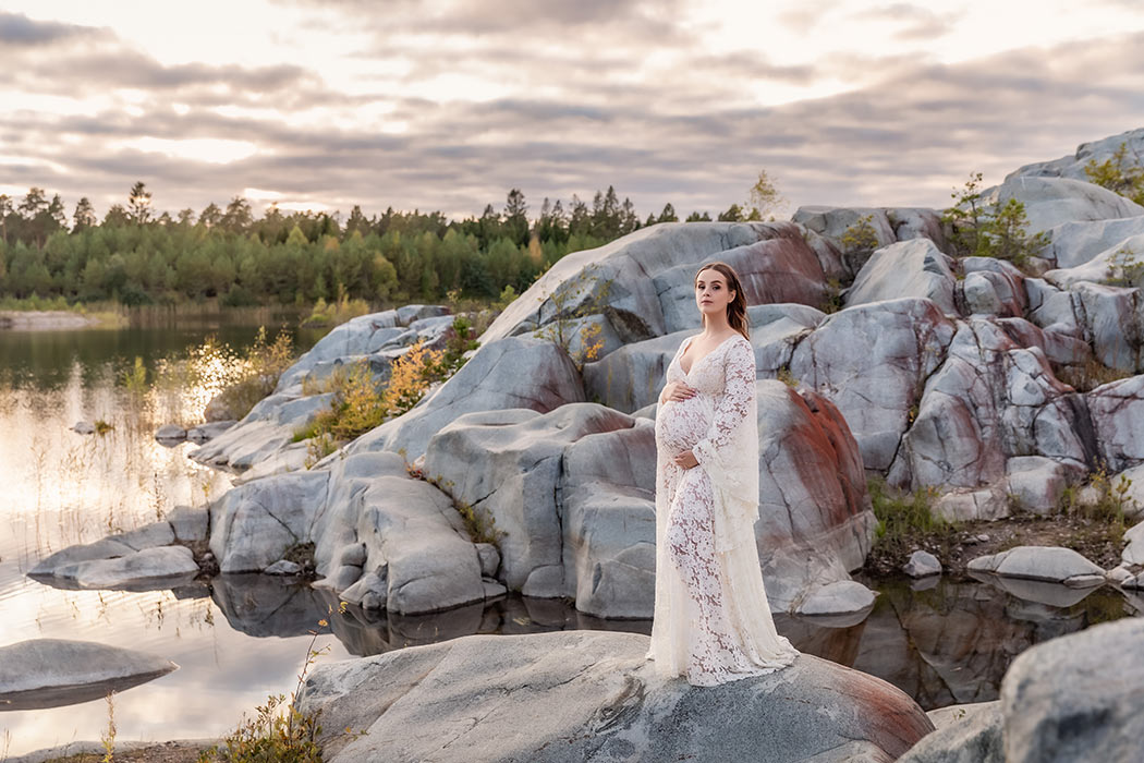 En kvinna i vit spetsklänning står i ett landskap av stora rundade klippor. Hon är gravid och tittar in i kameran. I vattnet blänker solen som håller på att gå ner bakom skogen i bakgrunden.