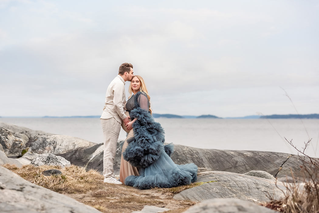 Gravidfotografering med ett par som står på klipporna med havet i bakgrunden. Den blivande pappan pussar den blivande mamman som tittar in i kameran.