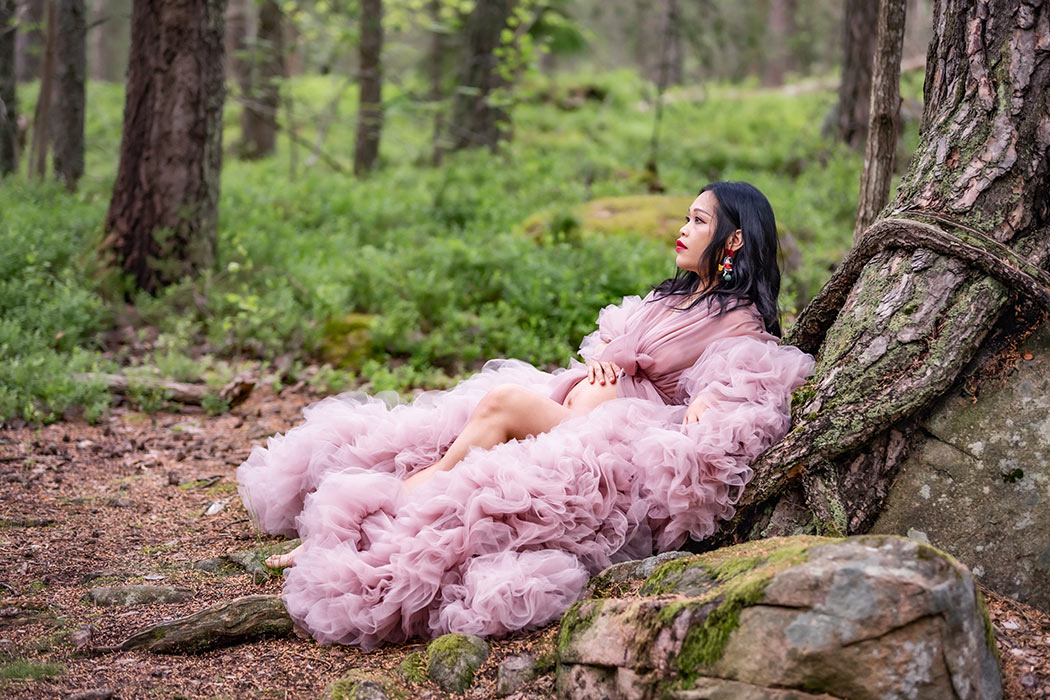 Gravidfotografering i skogen med en kvinna i rosa tyllklänning som lutar sig mot trädrötter.