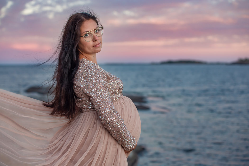 En gravid kvinna i glittrande rosa klänning tittar in i kameran. Hon står vid havet och himlen är vackert rosa.