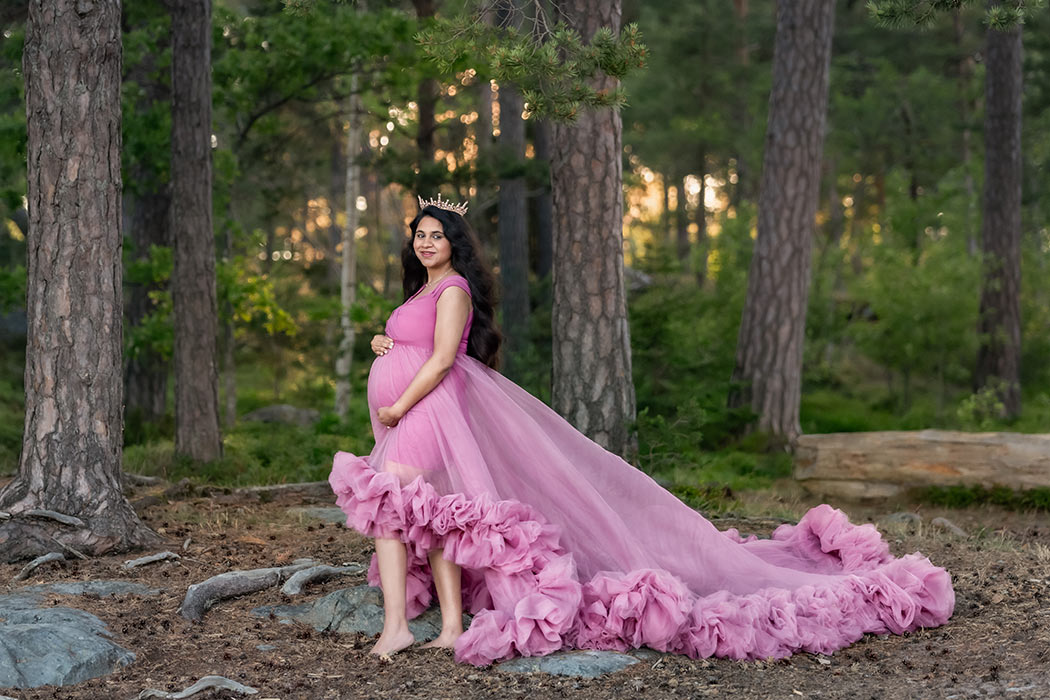 Gravidfotografering i en grönskande skog. Den gravida kvinnan har en rosa klänning med långt tyllsläp och en krona. Kvällsljuset letar sig in genom grenarna i bakgrunden.