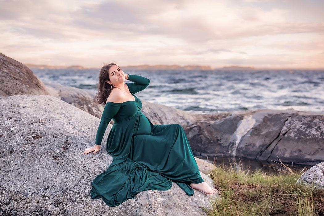 En vacker gravid kvinna i en grön klänning sitter på klipporna bredvid ett stormande hav. Himlen är molnbeklädd.