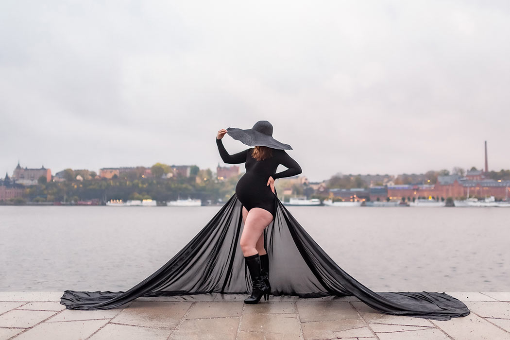 Gravidfotografering av en kvinna i svart polobody, långt svart släp och en gigantisk svart hatt. I bakgrunden syns vatten och en stadssiluett och det ligger regn i luften.
