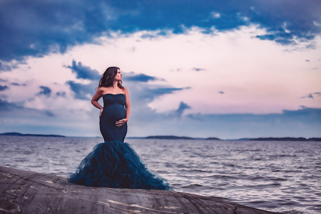 Gravidfotografering i september vid havet. Den gravida kvinnan har på sig en blå tyllklänning och står på klippor. I bakgrunden syns en blå och rosa himmel.