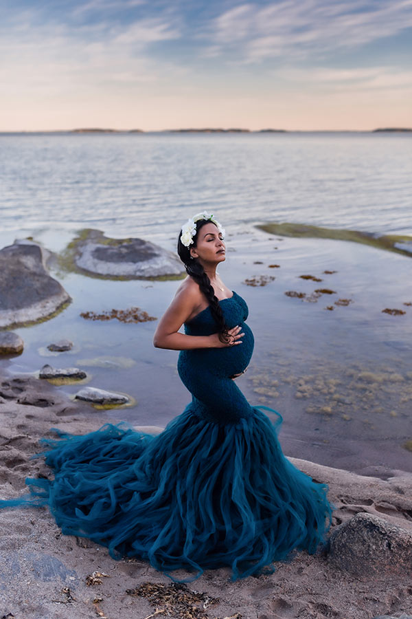 En gravid kvinna i blå tyllklänning står på en sandstrand. I bakgrunden syns stenar i vattnet och en vacker himmel.