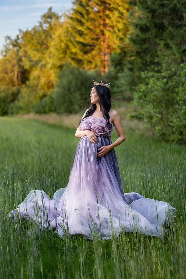 Gravidfotografering på en åker bredvid en grönskande skog. Den gravida kvinnanhar en vacker lila och rosa klänning och en krona.