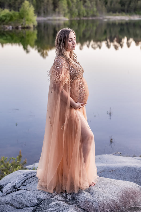 Stilla gravidfotografering vid en blank sjö. Den gravida kvinnan har en lyxig gravidklänning och i bakgrunden speglas träd i vattnet.