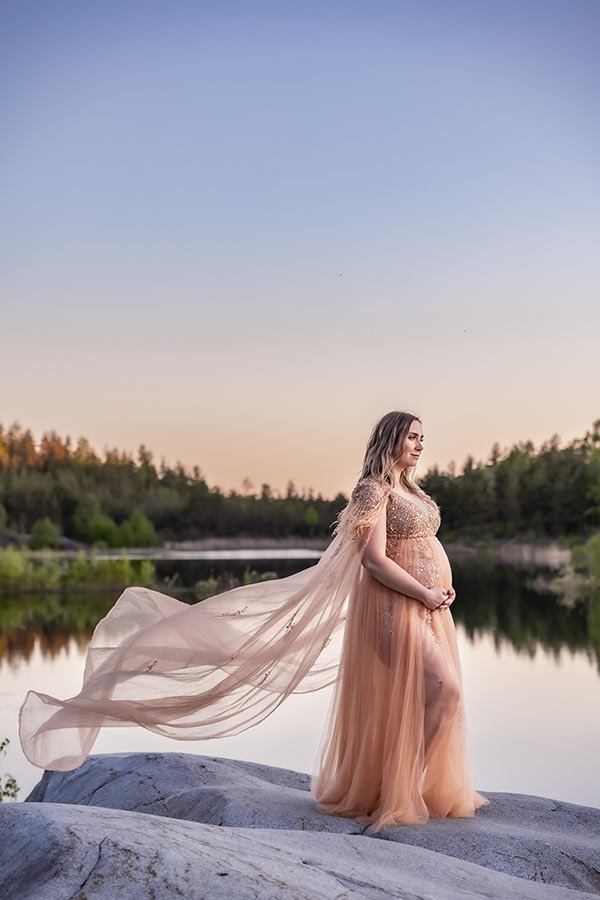 Gravidfotografering på klipporna vid en liten sjö. Den gravida kvinnan har på sig en persikofärgad lyxig gravidklänning med glitter och fjädrar och en tyllmantel som blåser i vinden.