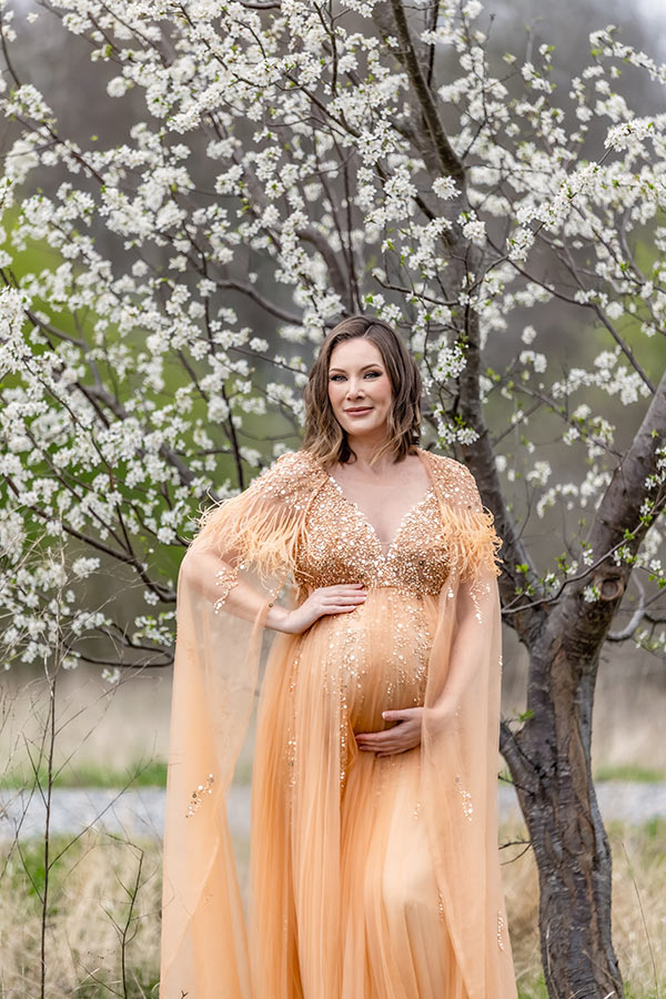 Gravidfotografering framför ett blomstrande träd. Den gravida kvinnan har en glittrande klänning och tittar in i kameran.
