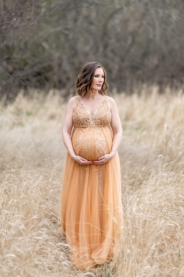 En gravid kvinna står på en äng med gulnat gräs. Hon har på sig en glittrande persikofärgad klänning.