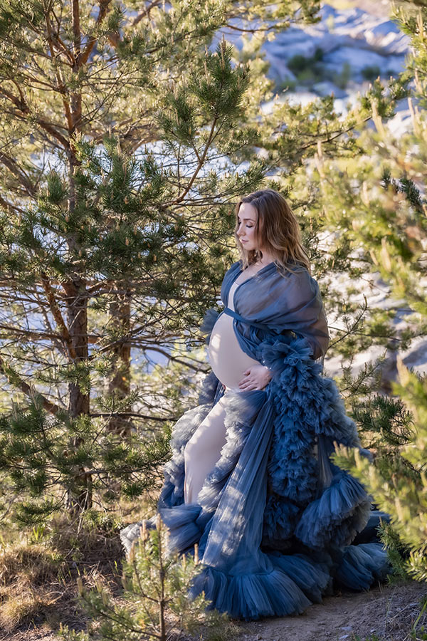 Gravidfotografering vid ett stilla vatten. Kvinnan står i en skogsdunge men i bakgrunden skymtar vattnet.