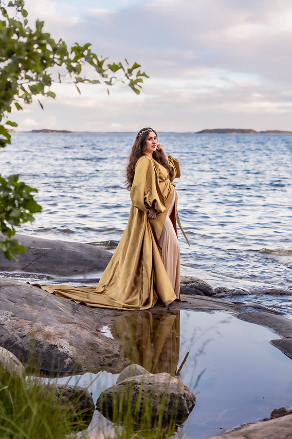 Gravidfotografering i skärgården bland klipporna. Den gravida kvinnan har en guldklänning och speglas i vattenpölen på klipporna. I bakgrunden syns havet.