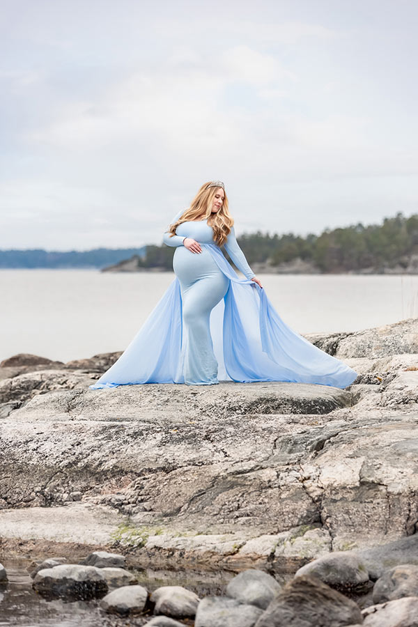 En gravid kvinna står på klipporna med en blå klänning.