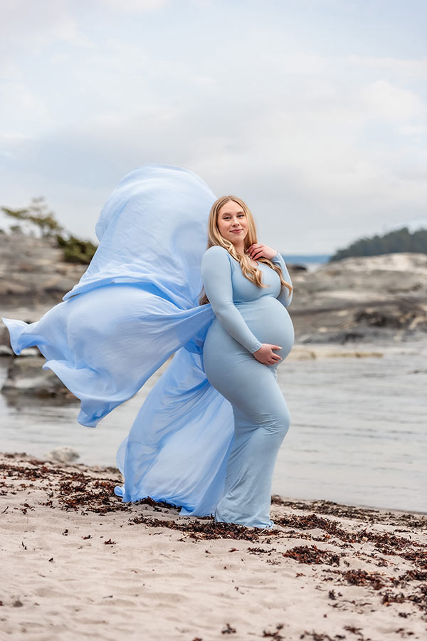 En gravid kvinna står på en sandstrand med en blå klänning med flygande släp.