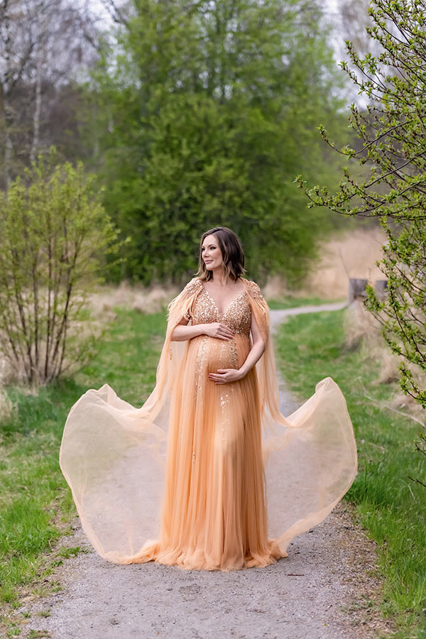 Gravidfotografering om våren bland träd och buskar som just börjat knoppas. Den gravida kvinnan har en glittrande persikofärgad klänning med en tunn tyllmantel som blåser i vinden.