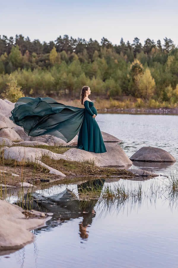 Gravidfotografering i ett stenbrott. En gravid kvinna i grön gravidklänning med långt släp står på några stora stenar vid vattnet. Det långa släpet flyger i vinden och kvinnan spegla i det stilla vattnet.