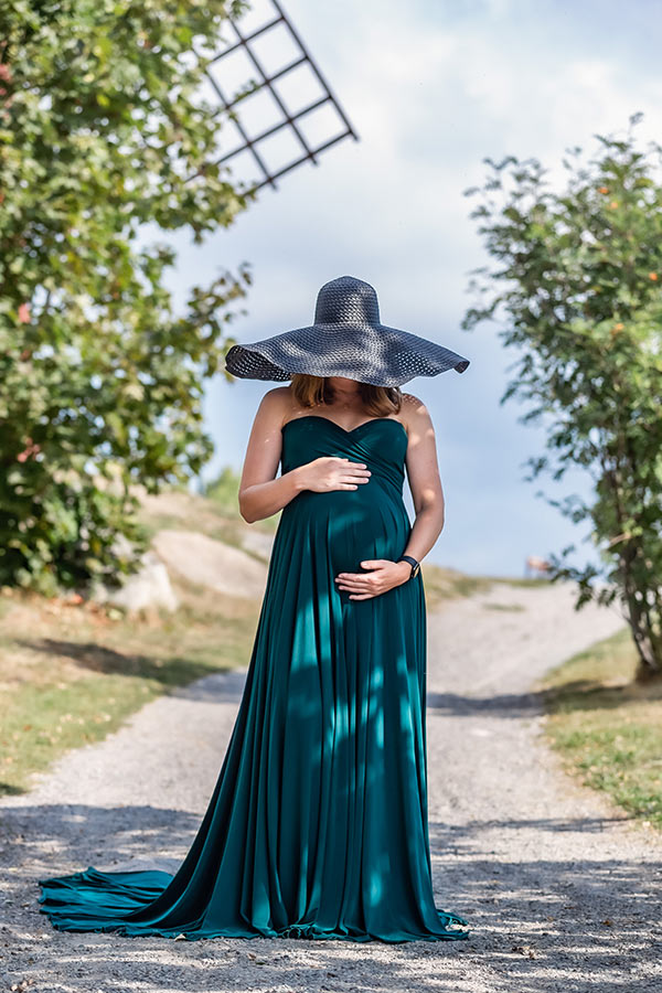 En gravid kvinna i grön klänning och svart hatt med stort brätte står på en grusstig som går uppför en kulle. Gången kantas av grönska.