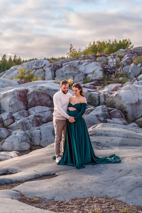 Gravidfotografering i ett stenbrott i solnedgången. Den gravida kvinnan har en vacker grön gravidklänning och står tillsammans med sin partner, den blivande pappan, och tittar ner på magen.