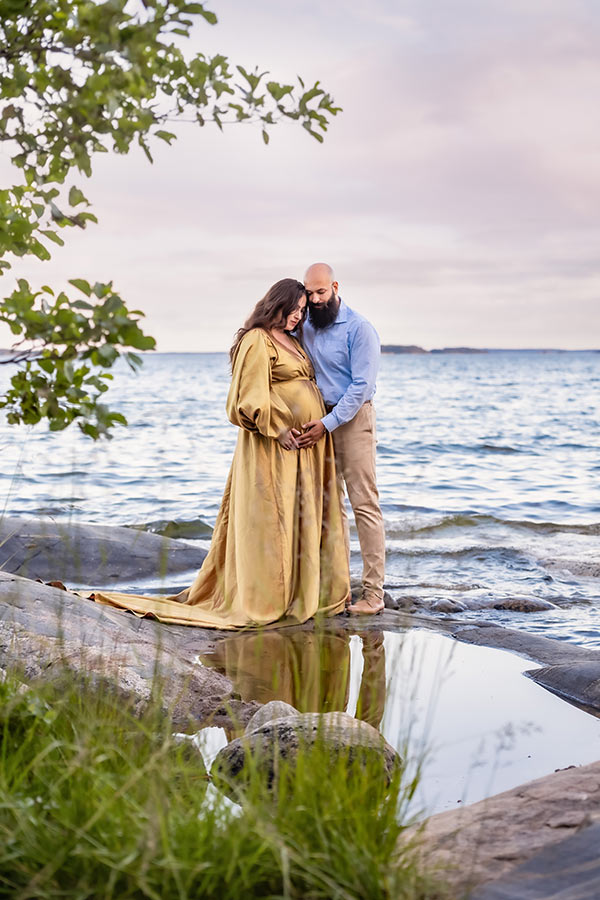 Gravidfotografering med ett par som står på klipporna i skärgården. I förgrunden syns gräs och spegelblank pöl och i bakgrunden syns ett vågigt hav.