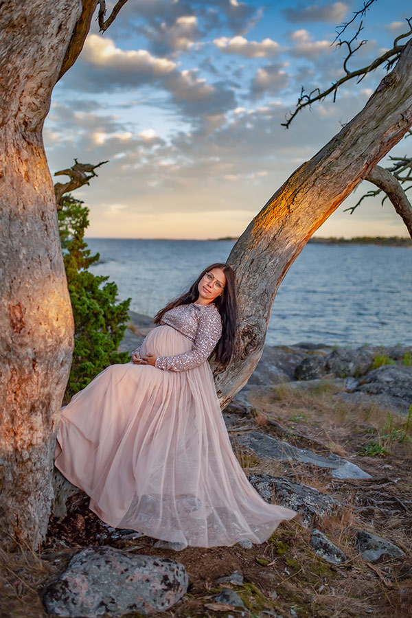Gravidfotografering på vintern på en stenstrand med vass. Den gravida kvinnan har på sig en grön klänning och tittar ut över havet. På vattnet ligger is och himlen är täckt av moln.