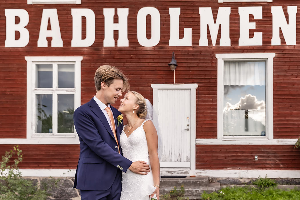 Ett vackert brudpar kramas och tittar på varandra. De står framför ett rött hus med vita knutar som har stora vita bokstäver där det står Badholmen.