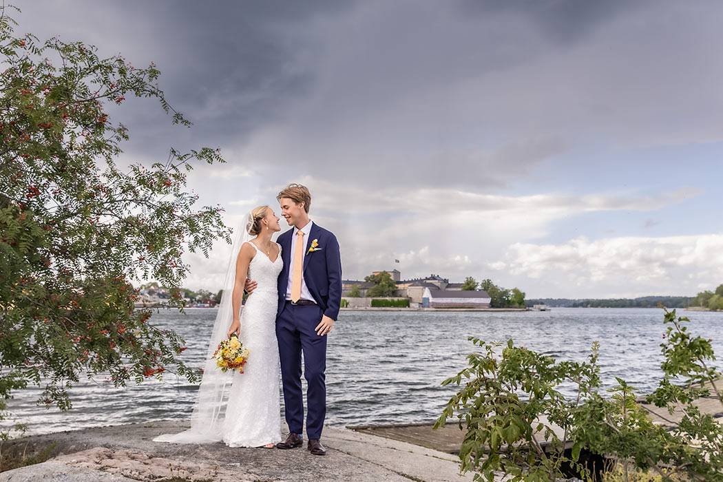 Bröllopsfotografering på skärgårdsön Badholmen. Paret står på en klippa och tittar leende på varandra. På andra sidan vattnet syns Vaxholm under mörka moln och regn.