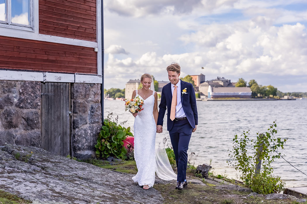 Bröllop en vacker sommardag på den lilla ön Badholmen utanför Vaxholm. Brudparet går hand i hand från bryggan och i bakgrunden syns Vaxholms kastell under stora moln.