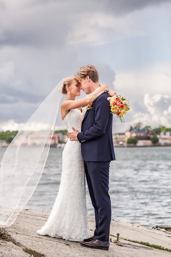 Bröllopsfotografering på skärgårdsön Badholmen. Paret står på en klippa och håller om varandra. På andra sidan vattnet syns Vaxholm.