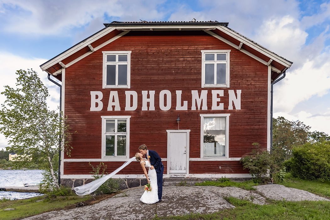 Ett vackert brudpar kysser varandra framför framför ett rött hus med vita knutar som har stora vita bokstäver där det står Badholmen. Brudens slöja flyger i vinden och runt huset syns grönskande sommar och hav.