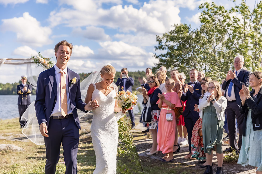 Glada och nygifta går brudparet från vigselplatsen vid havet medan bröllopsgästerna applåderar.