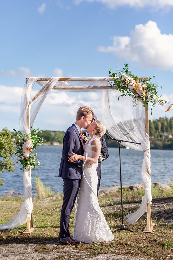 Bröllop på Badholmen utanför Vaxholm. Första kyssen som gifta under vigselbågen. Solen skiner stark där de står vid havet.