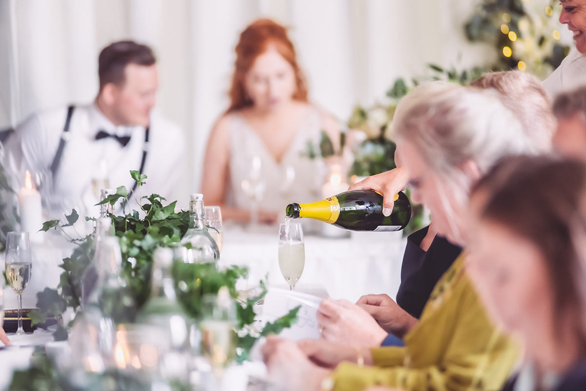 En bild från middagen på ett bröllop. Gästerna är ur fokus och en champagneflaska som häller upp mer bubbel i ett glas är i fokus.