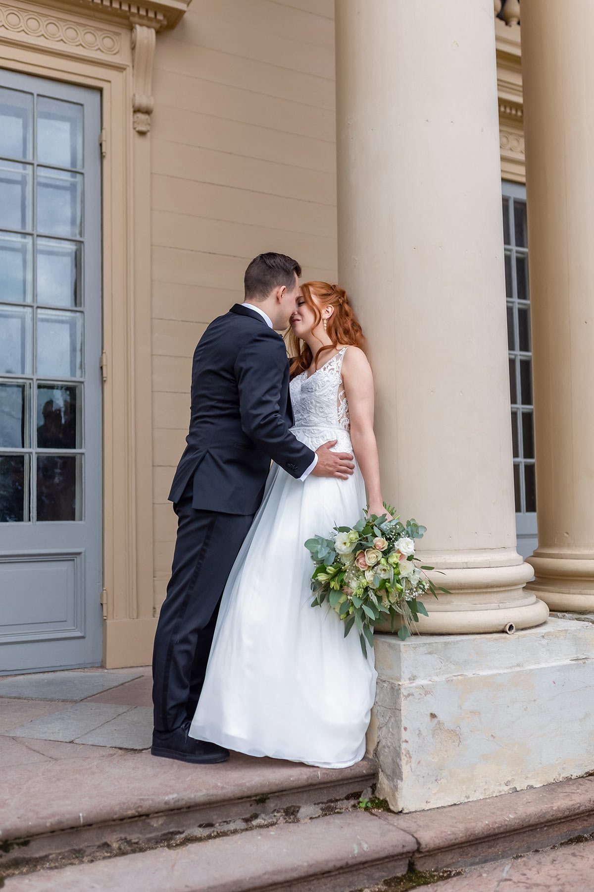Ett bröllopspar pussas vid en vacker byggnad. Bruden lutar sig mot en pelare