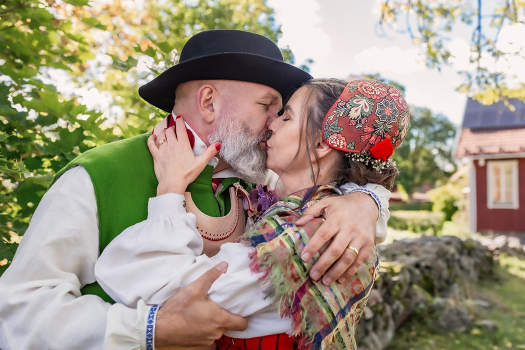 Ett brudpar i folkdräkt kysser varandra. I bakgrunden syns en stenmur, grönska och en faluröd stuga med vita knutar.