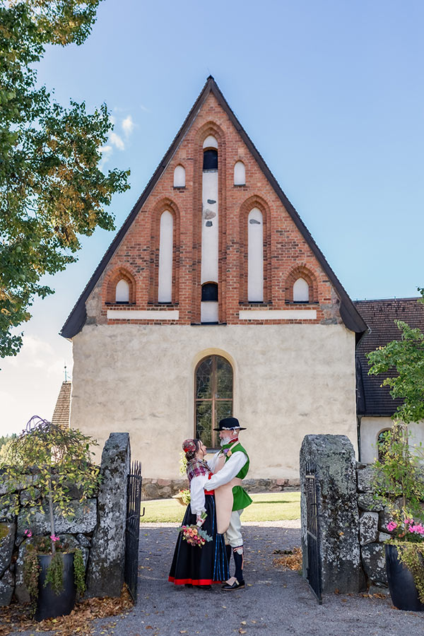 Bröllopsfotografering en vacker höstdag när solen skiner. Ett brudpar klädda i färgglada folkdräkter håller om varandra i grindöppningen till kyrkan. I bakgrunden syns grönska och S:t Stefans kyrka i Knivsta.