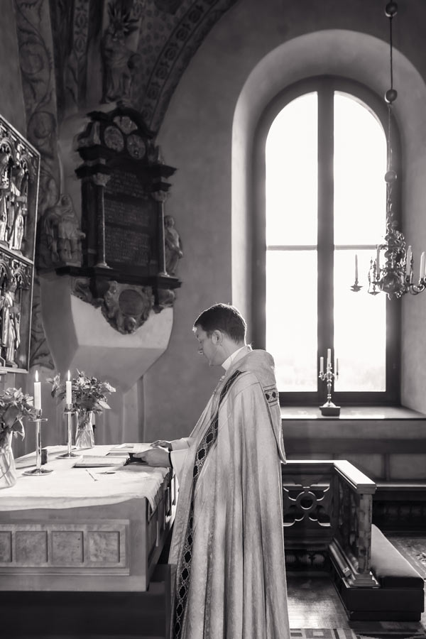 Svartvit bild på en präst som står framme i kyrkan. I bakgrunden syns ett stort vackert kyrkofönster.