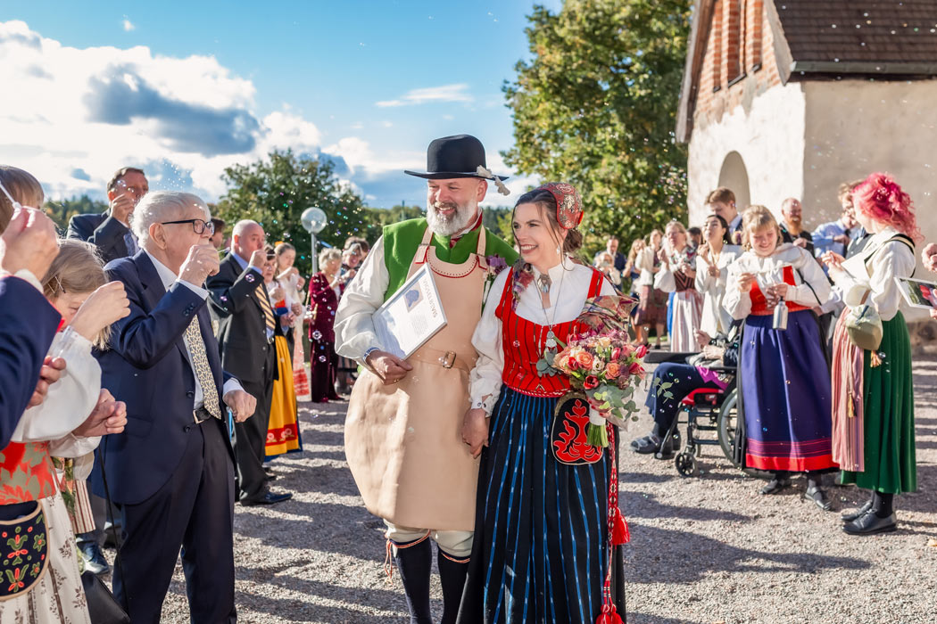 Ett brudpar klädda i folkdräkt går ut ur kyrkan medan deras nära och kära står runt dem och blåser sopbubblor. Det är vackert väder och solen skiner.