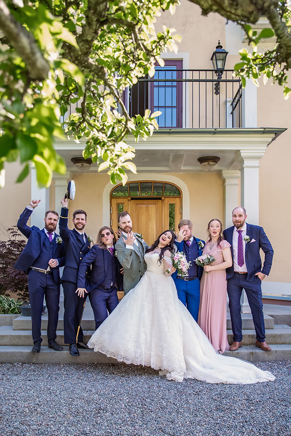 Brudparet och brudföljet fotograferas framför en av de vackra byggnaderna vid Södertuna Slott. I förgrunden syns grönskande grenar.